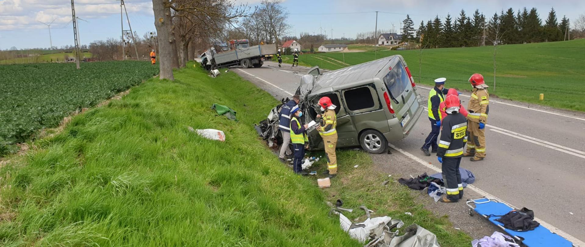 na drodze widać ratowników stojących przy wraku pojazdu po zderzeniu czołowym z innym pojazdem, który widać w oddali.