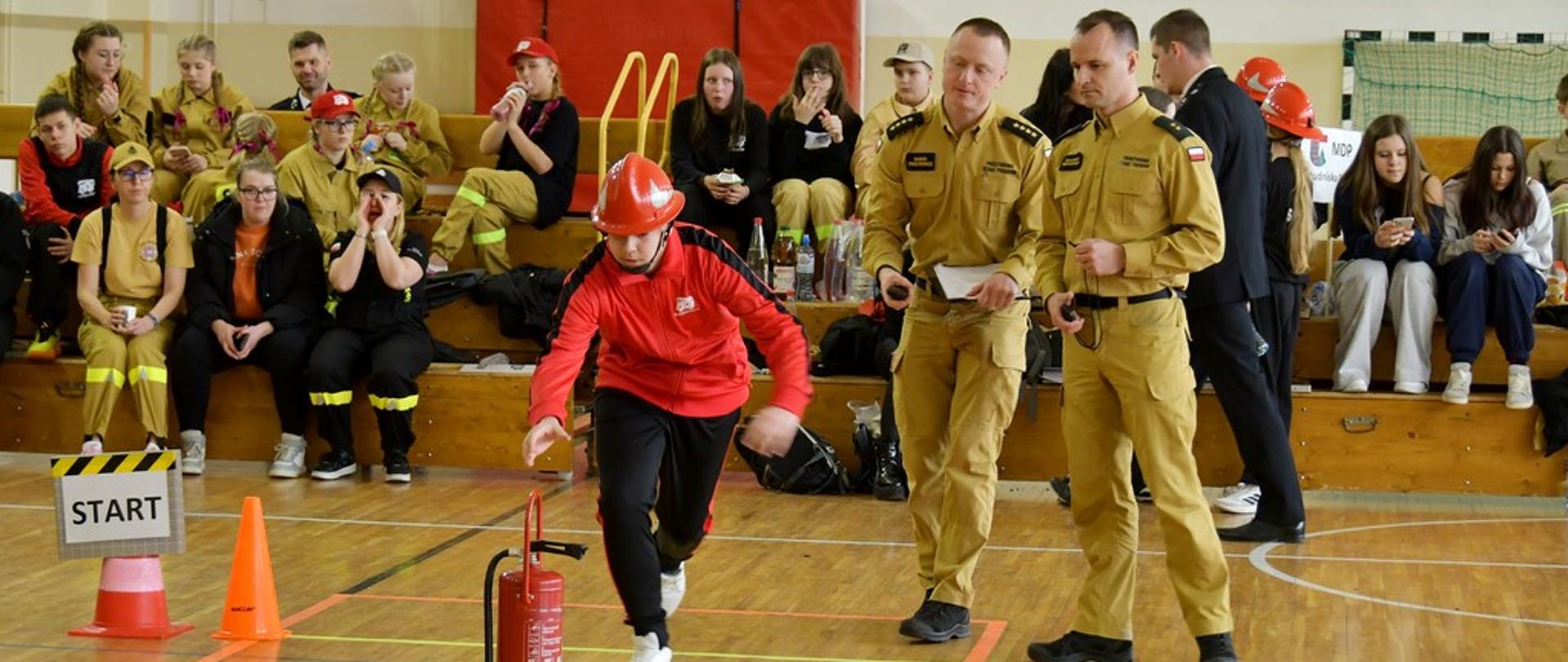 Na zdjęciu widać dziecko w czerwonej bluzie i czerwonym kasku, która biegnie po wyznaczonym torze w hali sportowej. Przed nią stoi gaśnica ustawiona na oznaczonej podstawie, a obok widoczny jest pachołek i tabliczka z napisem „START”. Po prawej stronie stoją dwaj funkcjonariusz PSP w piaskowych mundurach służbowych, obserwujący przebieg zadania i trzymający dokumenty. W tle na drewnianych ławkach siedzi grupa młodzieży, część z nich ma na sobie stroje strażackie i przygląda się sytuacji.