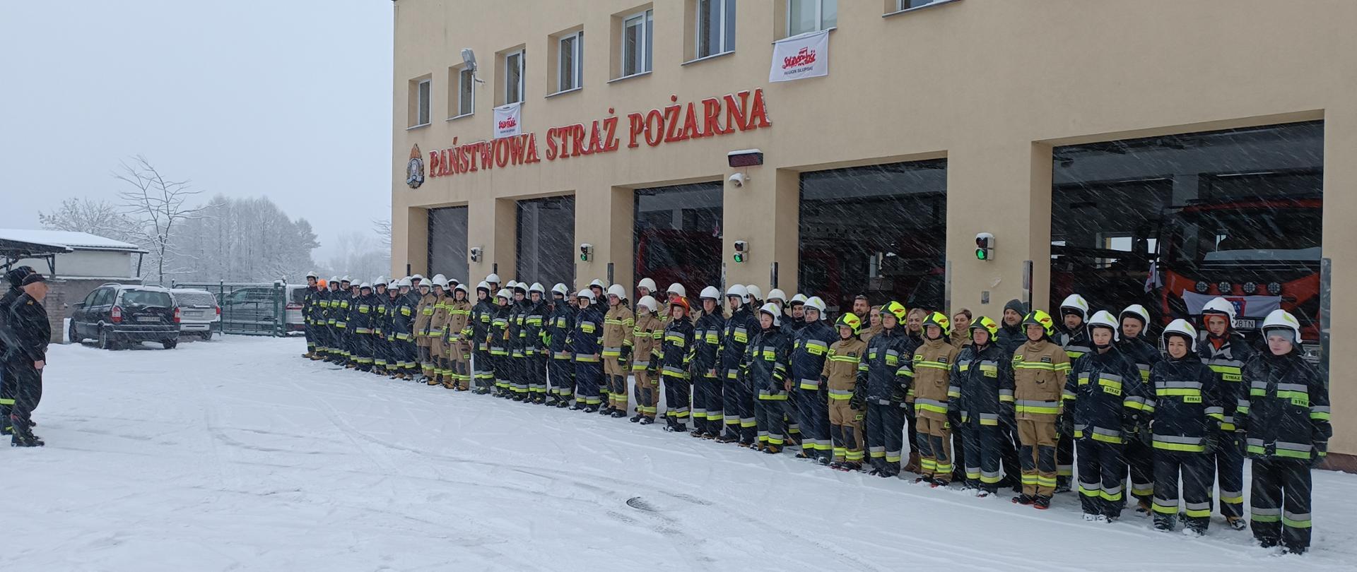 Zdjęcie przedstawi grupę strażaków ustanowionych w szyku na tle budynku straży pożarnej podczas zbiórki po zakończonym kursie strażaków ratowników.