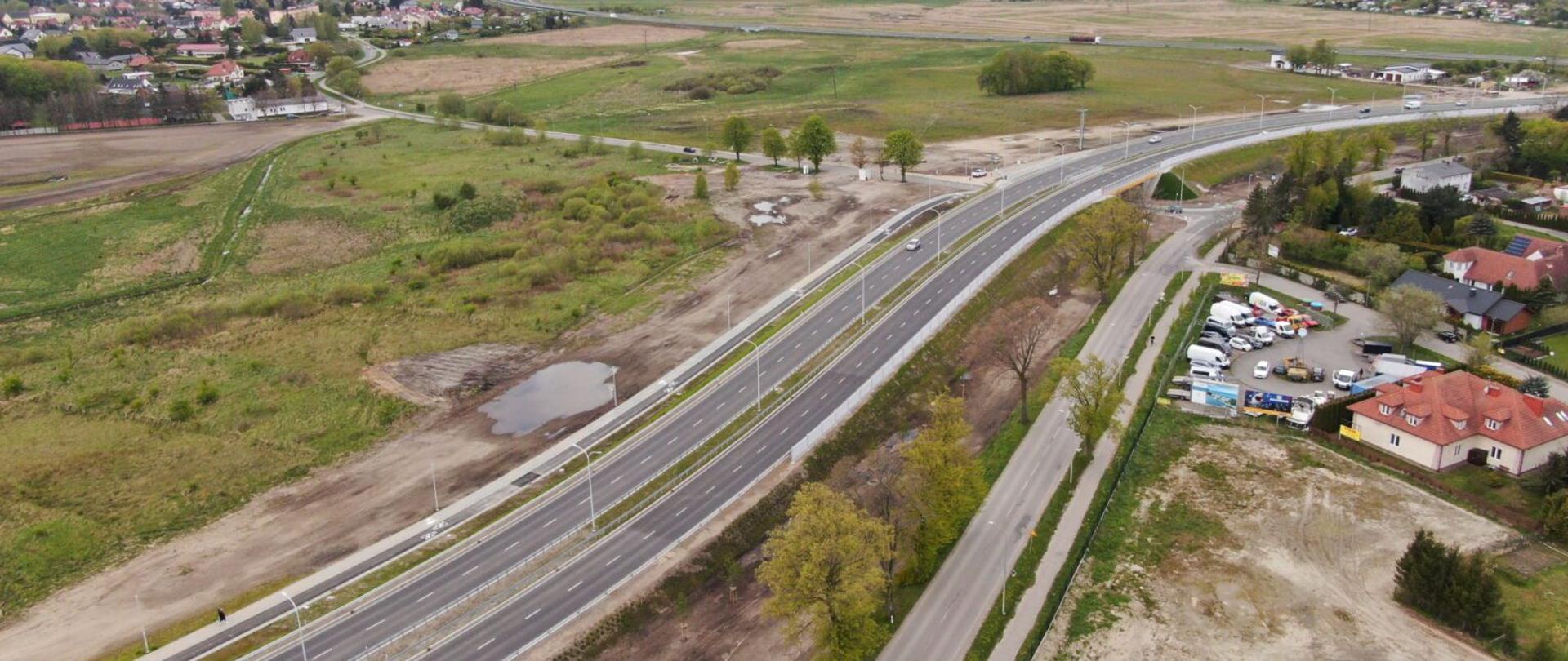 Zdjęcie lotnicze przedstawia drogę szybkiego ruchu biegnącą przez środek kadru, otoczoną zielonymi polami i zabudową. Po prawej stronie widoczne jest osiedle domów jednorodzinnych oraz parking z zaparkowanymi samochodami. W tle rozciągają się pola uprawne i dalsze zabudowania miejskie.