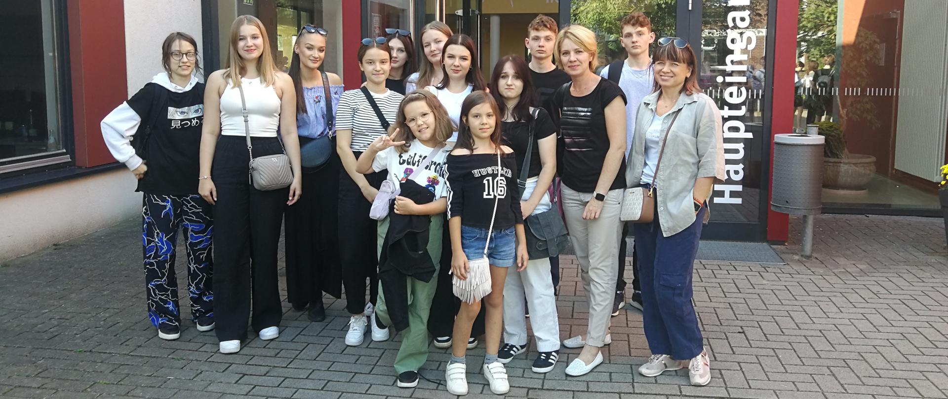 Zdjęcie przedstawia grupę uczniów i nauczycieli przed wejściem do budynku. Od lewej: Malwina Piwińska, Oliwia Bielecka, Amelia Pankiewicz, Gabriela Kościk, Kamila Wróblewska, Amelia Lewkowicz, Katarzyna Wiśniewska, Matylda Kędra, Lena Torba, Jagoda Piwińska, Mateusz Zieliński, Małgorzata Kawalec-Pyś, Radosław Zieliński, Iwona Grabowska