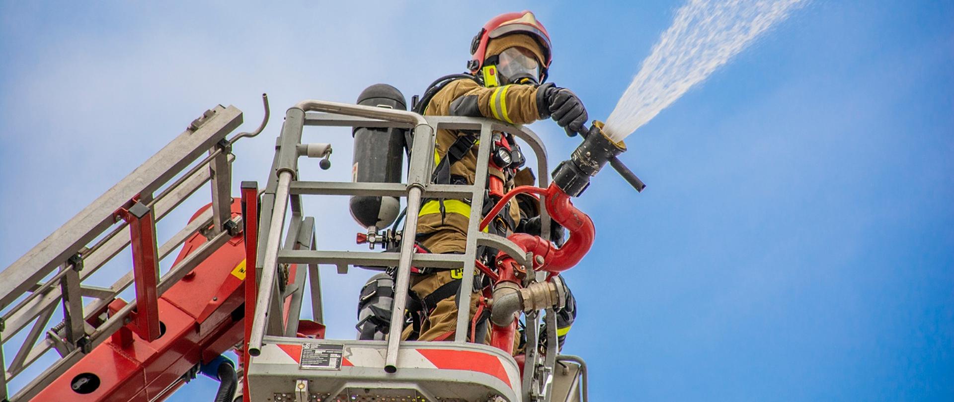 Na zdjęciu strażak w koszu podnośnika podający wodę na dach zakładu produkcyjnego.