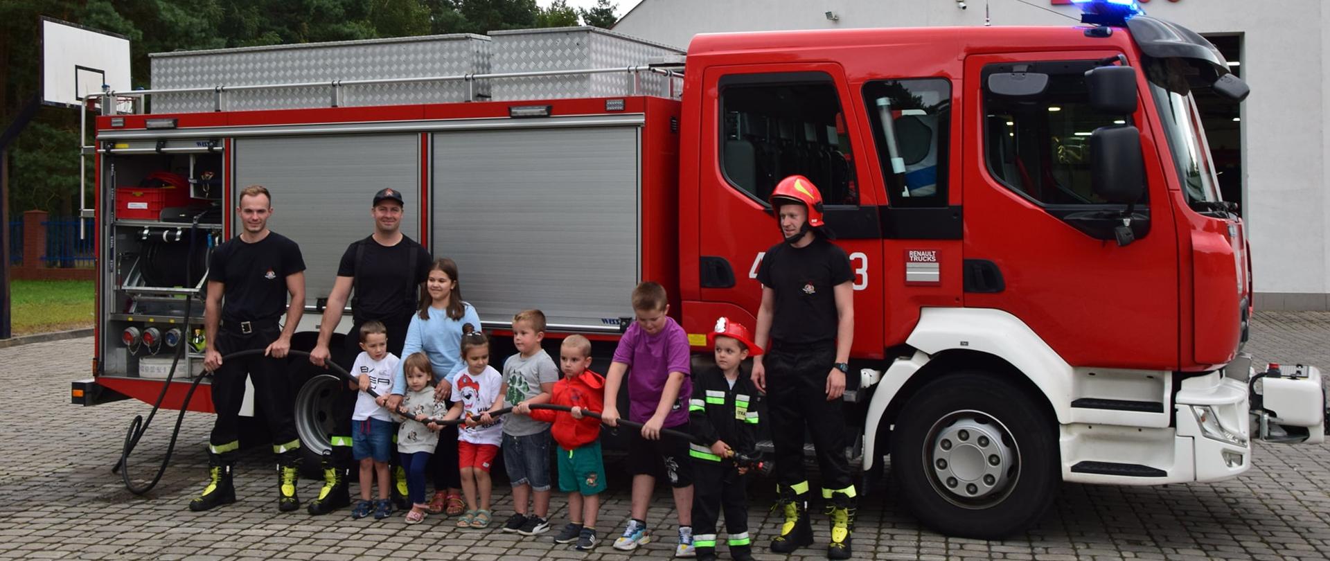 Na zdjęciu widać grupę dzieci ustawionych na tle wozu strażackiego, trzymających w dłoni węża zakończonego prądownicą. Z prawej strony stoi jeden strażak natomiast z lewej dwóch strażaków.