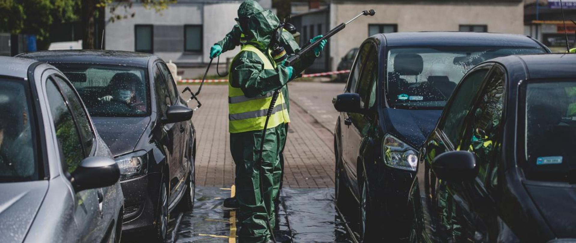 Wojska chemiczne odkażają pojazdy. Widoczni żołnierze w kombinezonach, którzy dokonują odkażania samochodów. W tyle parking i budynki.