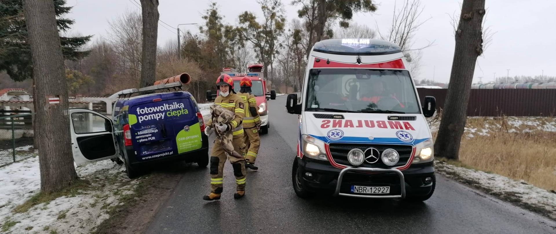 Na drodze ustawione pojazdy, Karetka, Straż Pożarna i z lewej strony auto które uderzyło w drzewo. Na drodze dwóch strażaków w czerwonych hełmach.