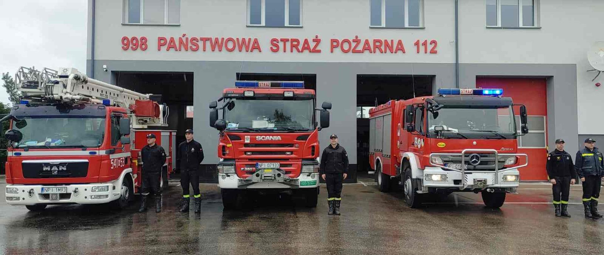Strażacy stojący na placu komendy obok wozów strażackich. Wozy z włączonymi światłami błyskowymi.