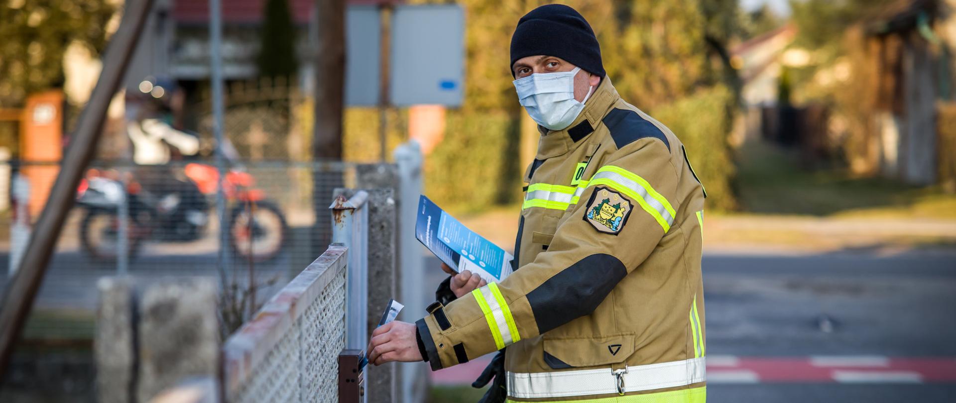 Zdjęcie przedstawia strażaka OSP wrzucającego ulotkę do skrzynki na listy umieszczonej na bramie posesji. Strażak w Ubraniu specjalnym w kolorze piaskowym, czarnej zimowej czapce oraz maseczce ochronnej. 