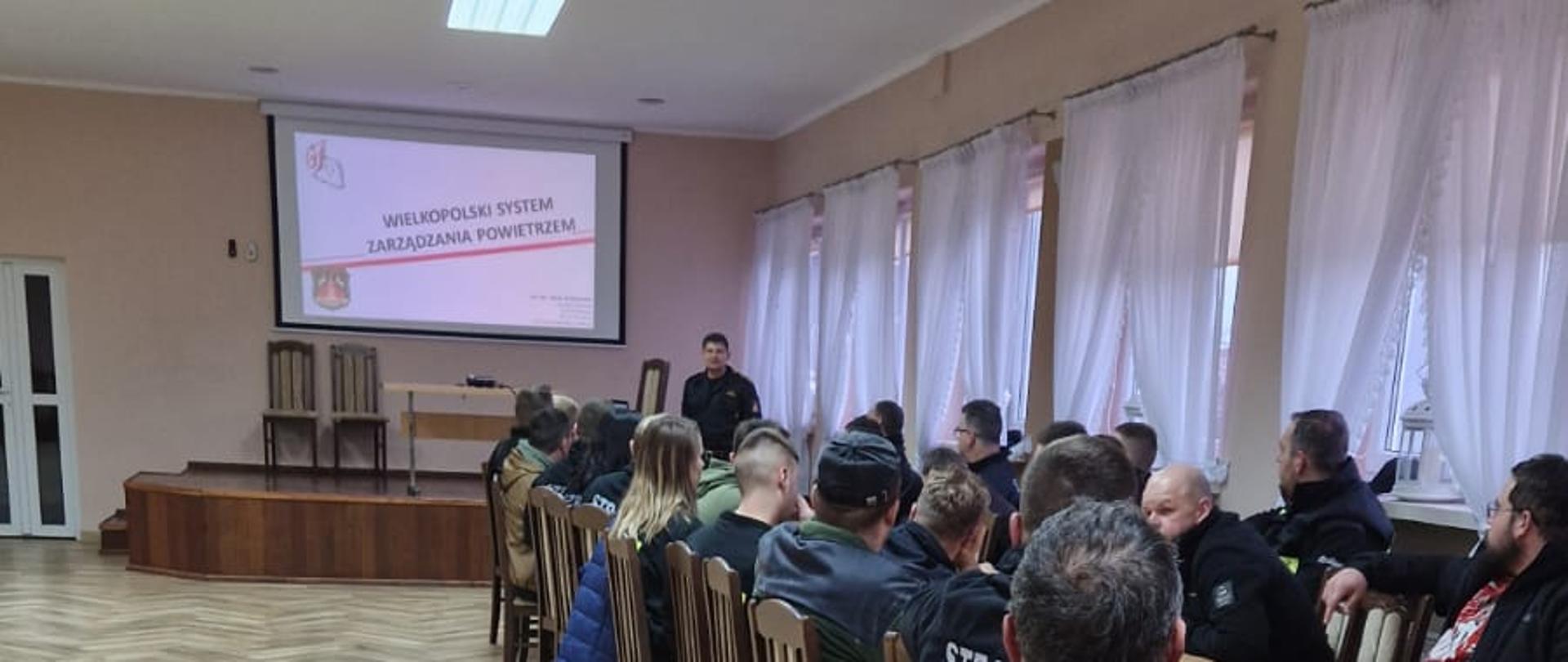 Zastępcę dowódcy JRG omawiającego prezentację na temat zarządzania powietrzem. Stoi on przy laptopie. Za nim na ścianie wyświetlana jest prezentacja. Przed nim przy stolikach siedzą druhowie.