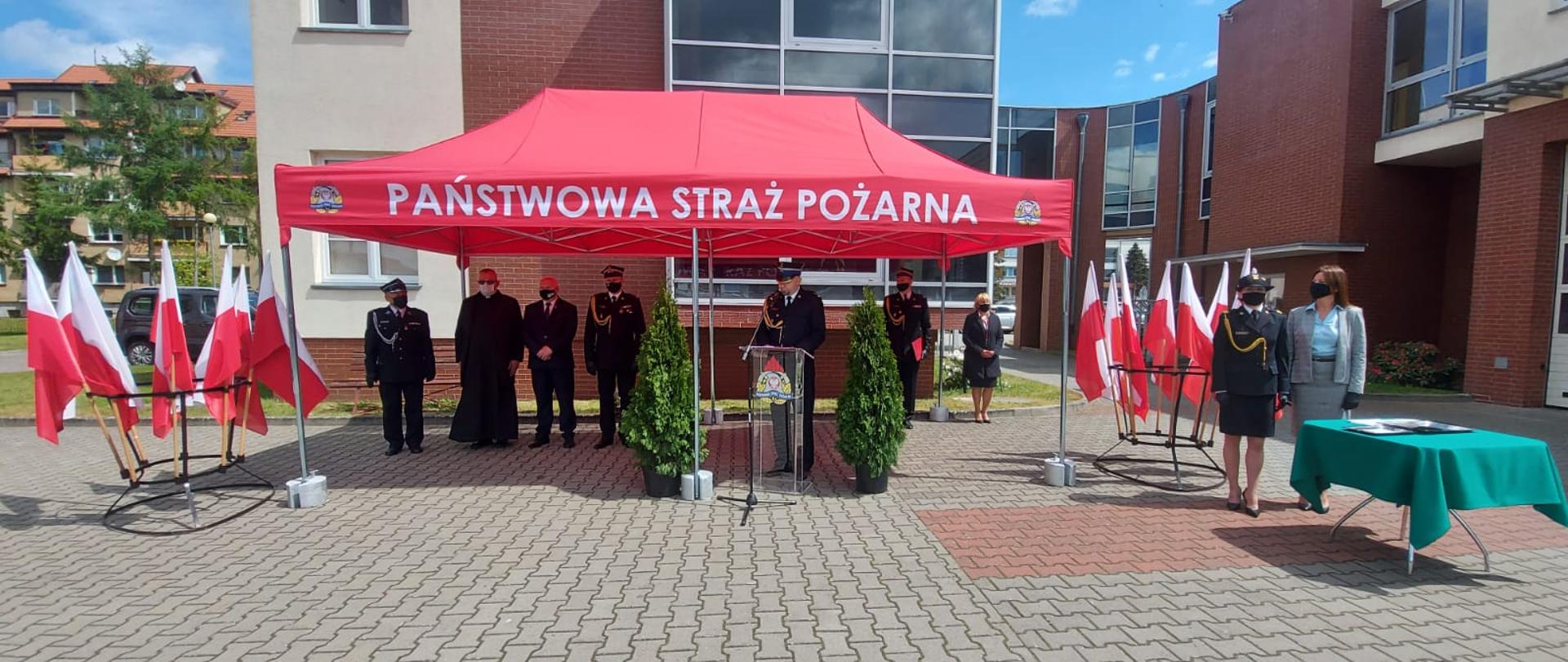 Zdjęcie przedstawia Strażaków oraz Gości stojących pod namiotem w czasie obchodów dnia strażaka. Po obu stronach namiotu postawione są stojaki z flagami Polski.w tle widać budynek komendy.