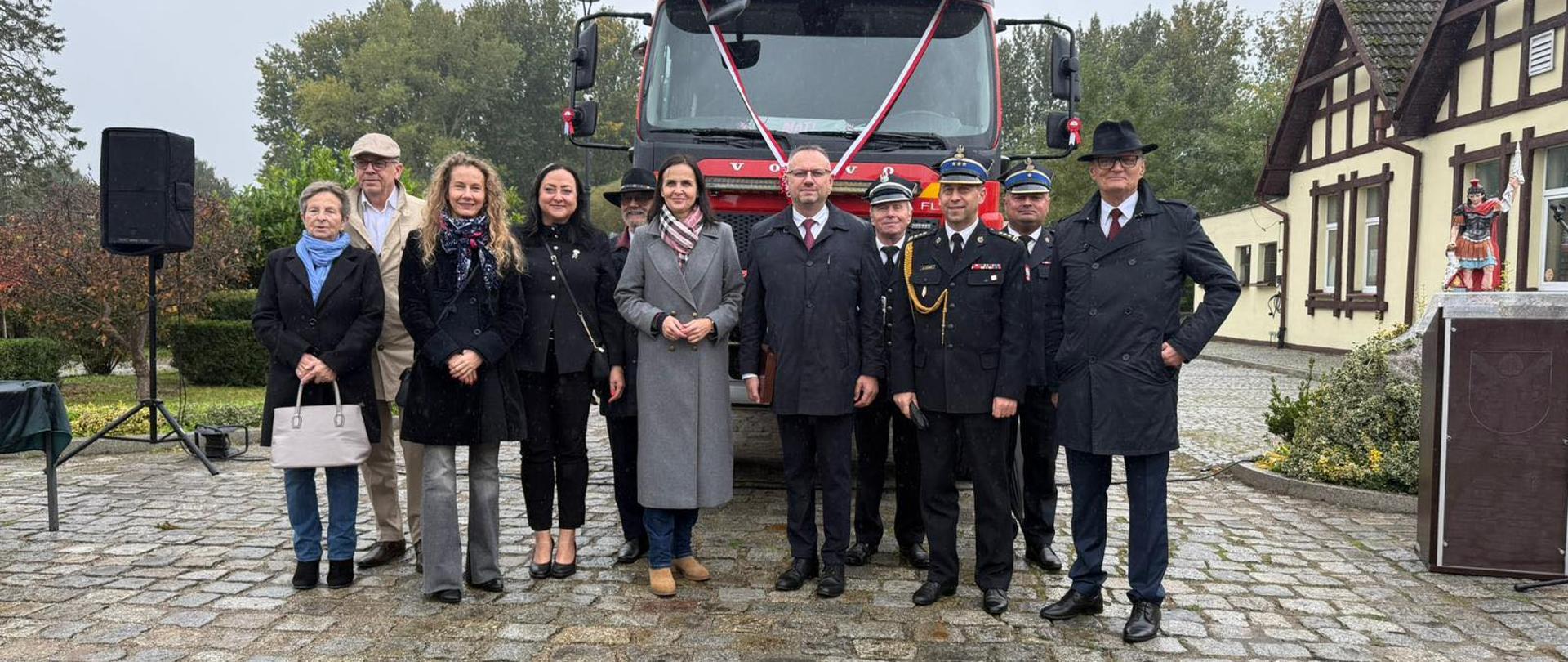 Grupa ludzi stoi przed czerwonym samochodem strażackim, wszyscy uśmiechają się i pozują do zdjęcia.