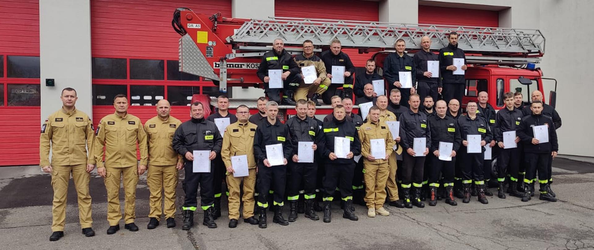 Grupa strażaków w ubraniach koszarowych koloru czarnego i piaskowego trzymająca zaświadczenia ukończenia szkolenia. Trzech funkcjonariuszy w ubraniach służbowych koloru piaskowego na tle samochodu bojowego i budynku komendy powiatowej.
