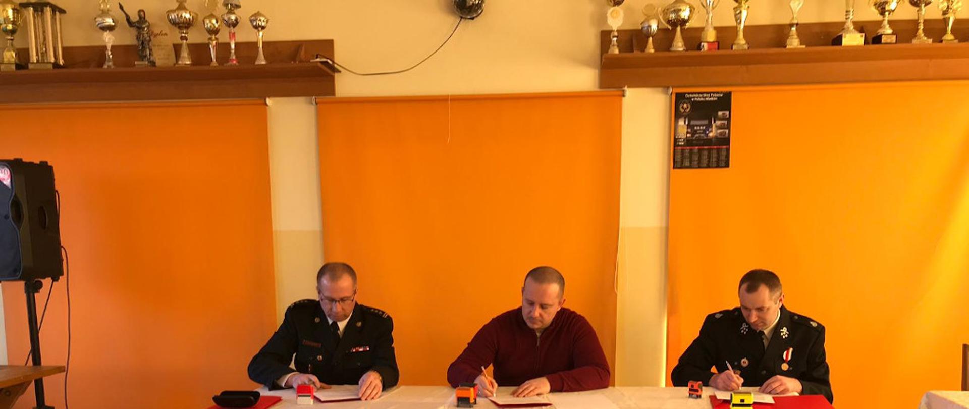 Na zdjęciu widnieje trzech mężczyzn siedzących przy stole w pomieszczeniu świetlicy. Cała trójka podpisje porozumienie dotyczące włączenie jednostki Ochotniczej Straży Pożarnej w Potoku Wielkim w struktury krajowego systemu ratowniczo-gaśniczego. Pierwszy z lewej to Komendant Powiatowy PSP w Jędrzejowie. Ubrany jest w galowy mundur. W środku znajduje się Burmistrz Miasta i Gminy Jędrzejów. Po lewej stronie stoi Prezes Ochotniczej Straży Pożarnej w Potoku Wielkim w umundurowaniu galowym OSP. W tle, na ścianie, znajdują się puchary ustawione na półce.