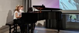 Małgorzata Nawrot i Krzysztof Wróblewski grający na cztery ręce na fortepianie. W tle na wysuniętym ekranie wyświetlony slajd z napisem Aleksander Tansman i podanymi datami urodzin i śmierci oraz zdjęcie biało-czarne patrona szkoły. 