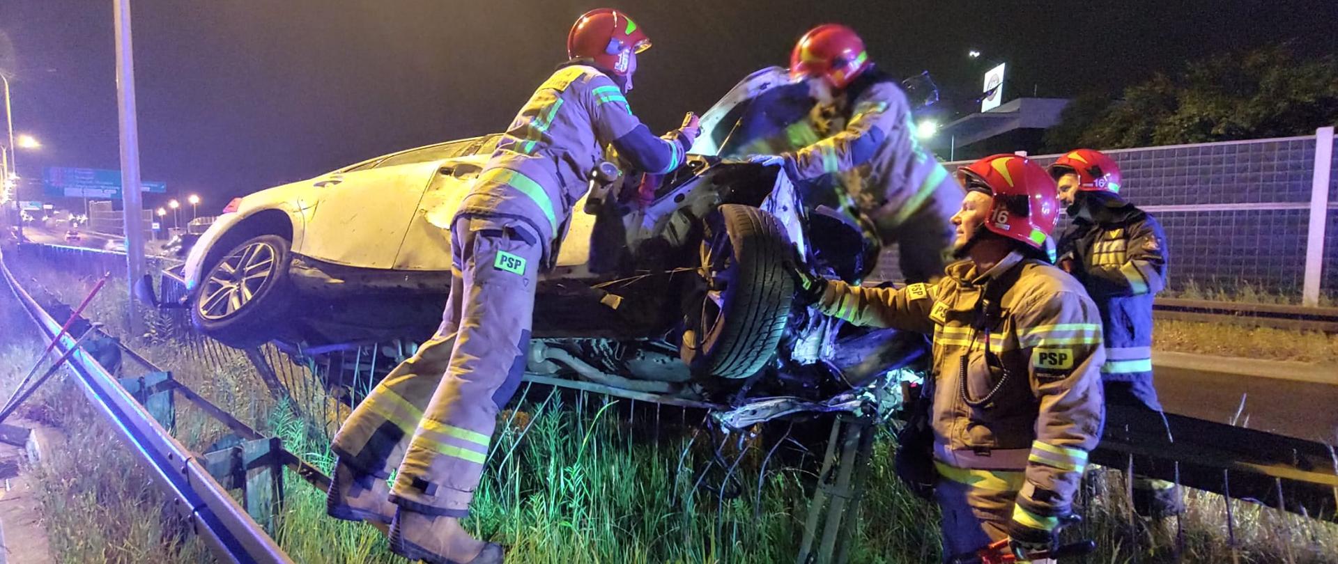 Na zdjęciu widoczni są strażacy pracujący przy zniszczonym aucie