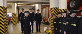 Na zdjęciu widać funkcjonariuszy PSP w umundurowaniu koszarowym, specjalnym oraz wyjściowym. Zdjęcie w pomieszczeniach komendy. Oświetlenie sztuczne. Podłoże z płytek.