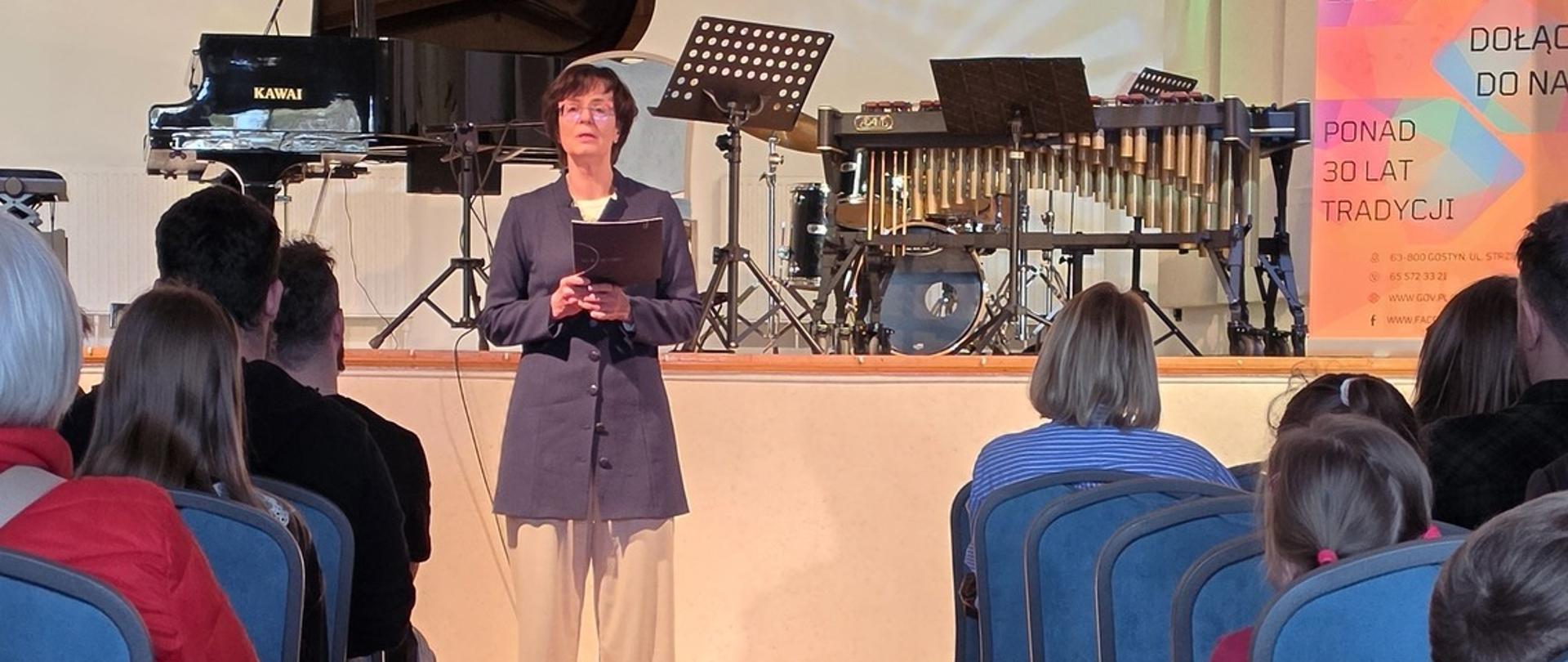 Zdjęcie przedstawia salę koncertową. Na sali siedzą uczestnicy wydarzenia. Na środku stoi pani Dyrektor i zabiera głos. Po prawej stronie znajduje się kolorowy rollup reklamujący szkołę. 