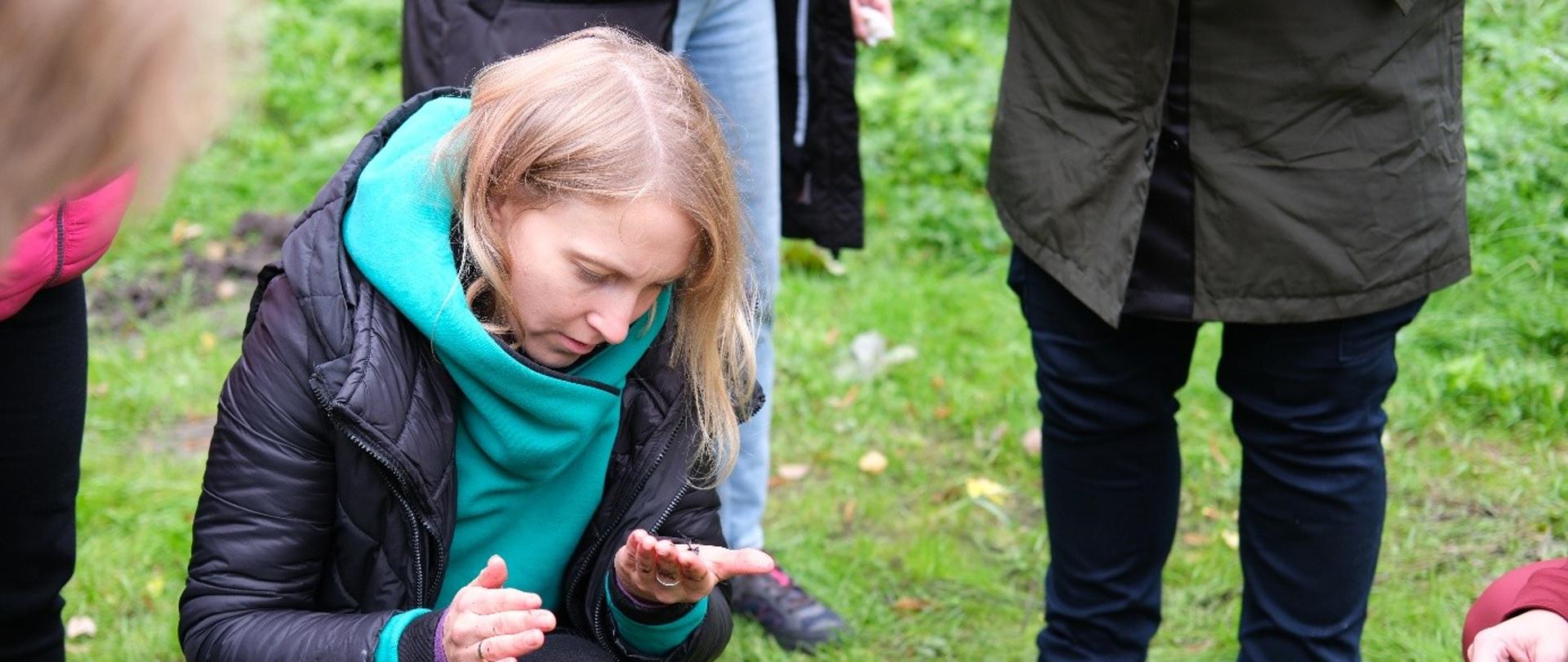 na zdjęciu widnieje uczestniczka warsztatów terenowej edukacji. Kobieta klęczy na trawie i ogląda na swojej dłoni małego owada. 
