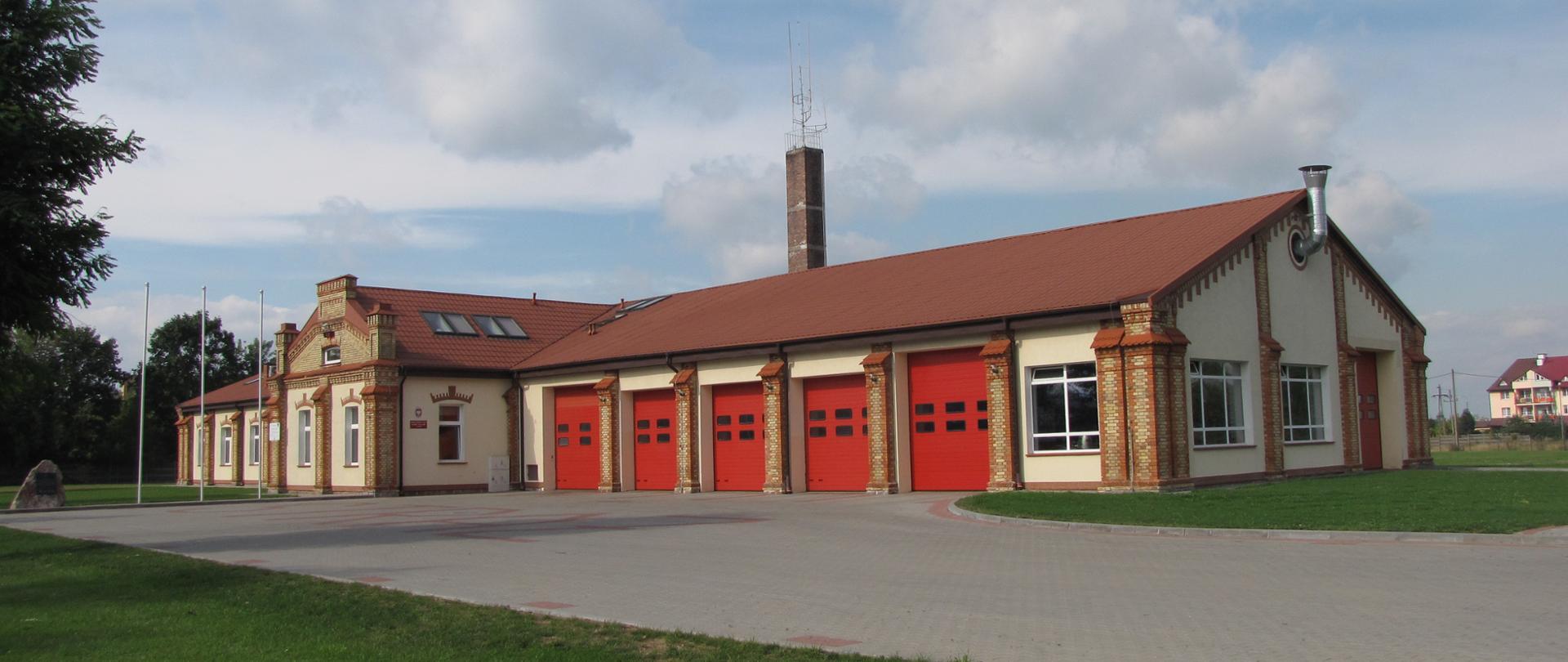 Na zdjęciu budynek Komendy Powiatowej Państwowej Straży Pożarnej w Grajewie. Widoczne czerwone bramy garażowe, budynek w kolorze jasnym piaskowym, dach brązowy, narożniki budynku z czerwonej i żółtej cegły. Z lewej strony zdjęcia widoczny kawałek drzewa liściastego.