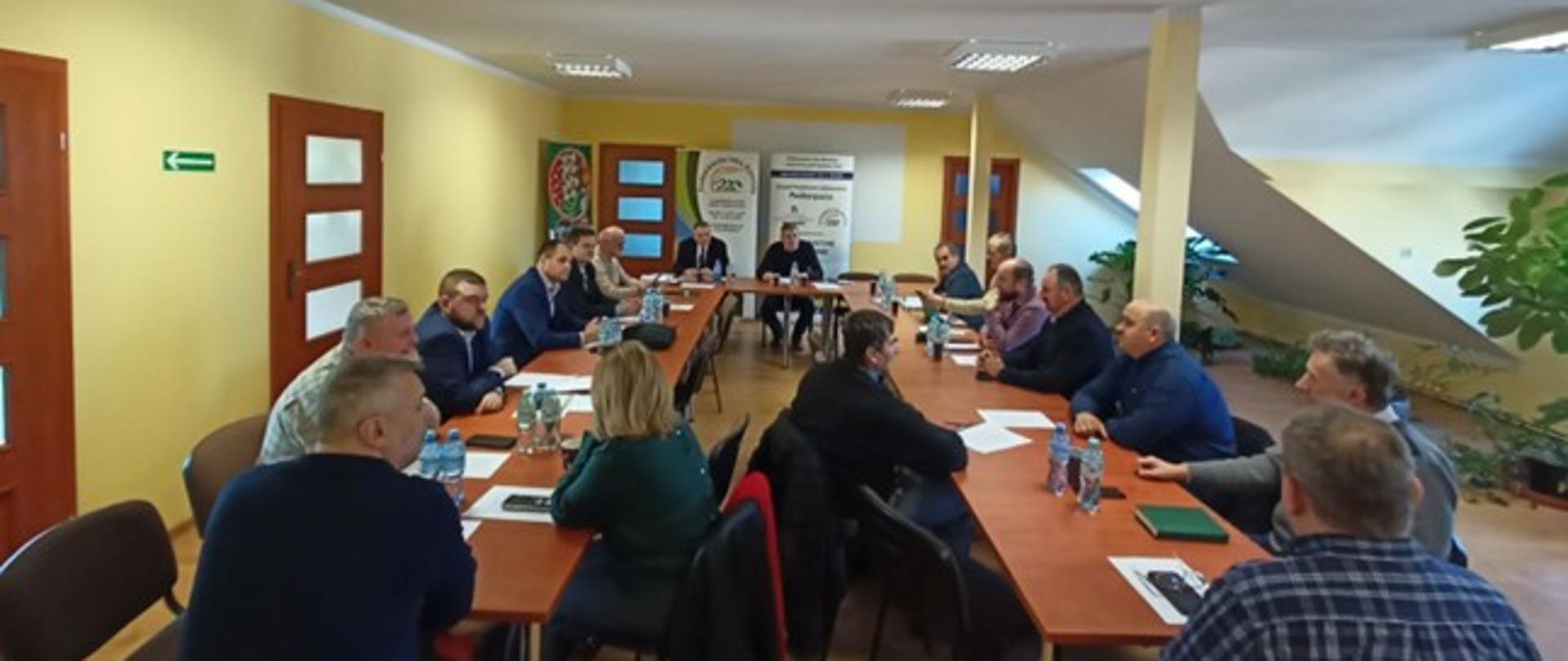 Zebranie grupy kilkunastu osób. Uczestnicy siedzą przy stołach ułożonych w kształcie litery U. Ściany pomieszczenia są żółte.