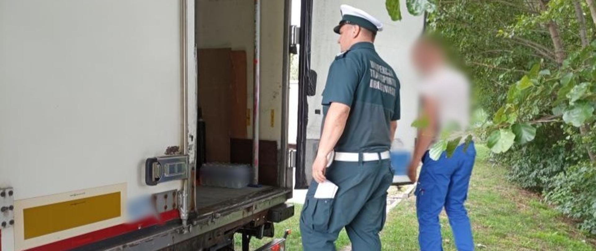 Umundurowany inspektor kujawsko-pomorskiej ITD stoi obok otwartych drzwi naczepy-chłodni i patrzy na ładunek przewożony w pojeździe. Po jego prawej stronie stoi kierowca kontrolowanej ciężarówki.