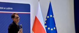 Na zdjęciu Szefowa służby cywilnej podczas spotkania w Lubelskim Urzędzie Wojewódzkim: stoi z mikrofonem przy mównicy, w tle flaga Polski i UE