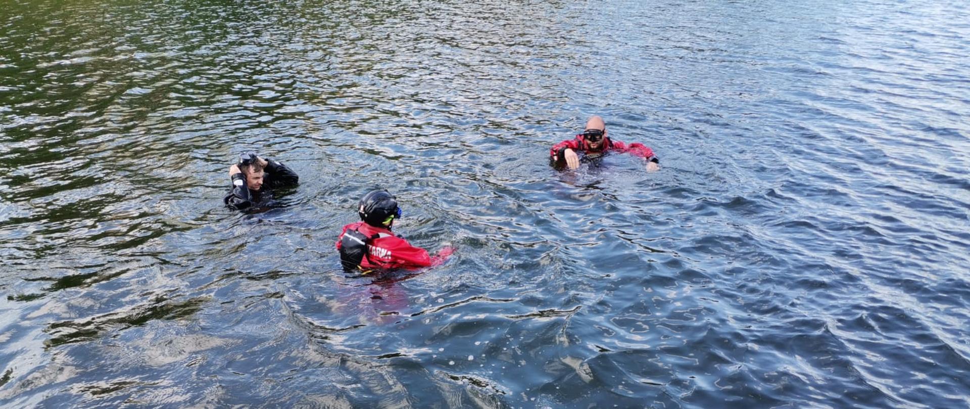 Trzech strażaków ubranych w ubrania do nurkowania zanurzonych w wodzie, na pierwszym planie na pomoście leży butla oraz pozostały sprzęt do nurkowania 