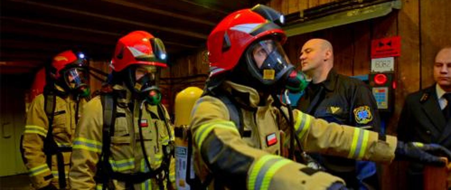 Trzej strażacy w umundurowaniu bojowym gotowi do startu w turnieju barbórkowym. W tle dwaj sędziowie.