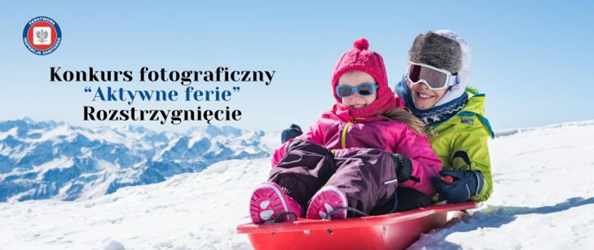 W zimowej scenerii dwoje dzieci (dziewczynka i chłopiec) zjeżdżających na czerwonych plastikowych sankach. Dziewczynka ubrana w kombinezon zimowy, czapkę i szalik w odcieniach różu. Chłopiec ubrany w odzież zimową w odcieniach zieleni w białej futrzanej czapce na głowie. Oboje dzieci mają założone okulary ochronne. Po lewej stronie napis konkurs fotograficzny "Aktywne ferie" - Rozstrzygnięcie. W lewym górnym rogu okrągłe logo Państwowej Inspekcji Sanitarnej przedstawiające czarnego orła na białym kwadratowym tle, które otoczone jest czerwonym wypełnieniem. Zewnętrzną część logo otacza granatowy okrąg z napisem Państwowa Inspekcja Sanitarna w kolorze białym.