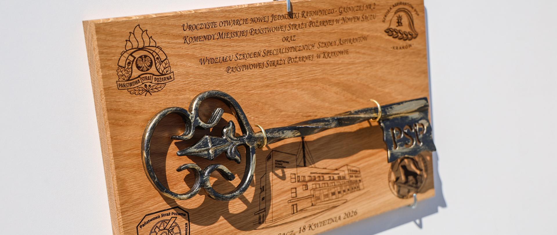 Drewniana tablica pamiątkowa z wygrawerowaną datą 18 kwietnia 2026 r. Na desce przymocowany jest duży, ozdobny, kuty klucz z napisem „PSP” na piórze. W tle widoczny grawer nowego budynku oraz logotypy Szkoły Aspirantów i Państwowej Straży Pożarnej.
