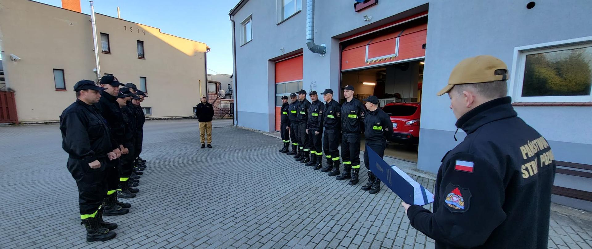 Strażacy w ubraniach koszarowych stoją w szeregu przed budynkiem Komendy Powiatowej Państwowej Straży Pożarnej w Sępólnie Krajeńskim, w tle garaże Jednostki Ratowniczo-Gaśniczej, na środku dowódca Jednostki Ratowniczo- Gaśniczej młodszy brygadier Robert Liss oraz Komendant Powiatowy starszy brygadier Maciej Suchomski w ubraniach służbowych 