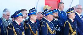 Grupa oficerów Państwowej Straży Pożarnej oraz zaproszonych gości stojących w szeregu podczas uroczystego apelu. Na pierwszym planie widoczni są czterej strażacy w ciemnych mundurach wyjściowych, czapkach rogatywkach ze złotymi okuciami, złotych sznurach galowych i czarnych rękawiczkach. W tle za nimi stoją cywile w garniturach oraz mężczyzna w zielonym mundurze leśnika.