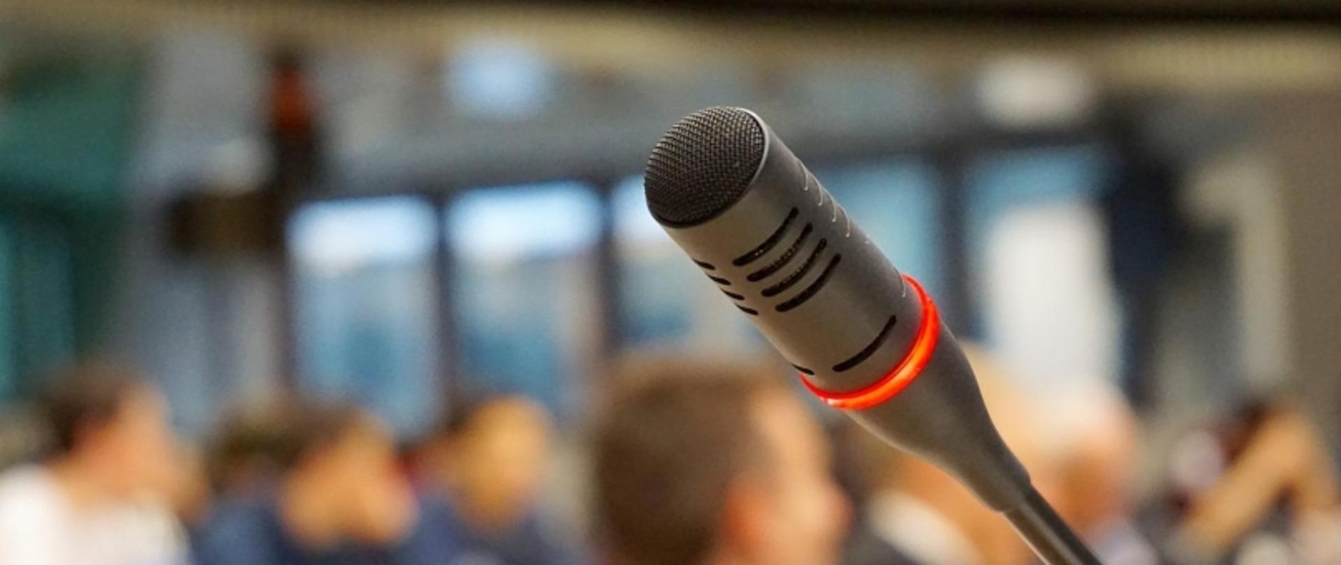 Zbliżenie na włączony mikrofon z czerwoną obwódką; w tle niewyraźne postacie uczestników spotkania.