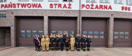 To zdjęcie zrobiono na zewnątrz, przed budynkiem straży pożarnej. Duża grupa strażaków stoi ustawiona w szeregu, pozując do zdjęcia. W tle widać budynek z czerwonej cegły z trzema dużymi, brązowymi bramami garażowymi. Nad bramami widnieje czerwony napis: "PAŃSTWOWA STRAŻ POŻARNA 998". Strażacy noszą dwa rodzaje mundurów: czarne, galowe mundury oraz mundury polowe w kolorze khaki i w czarnym kolorze.