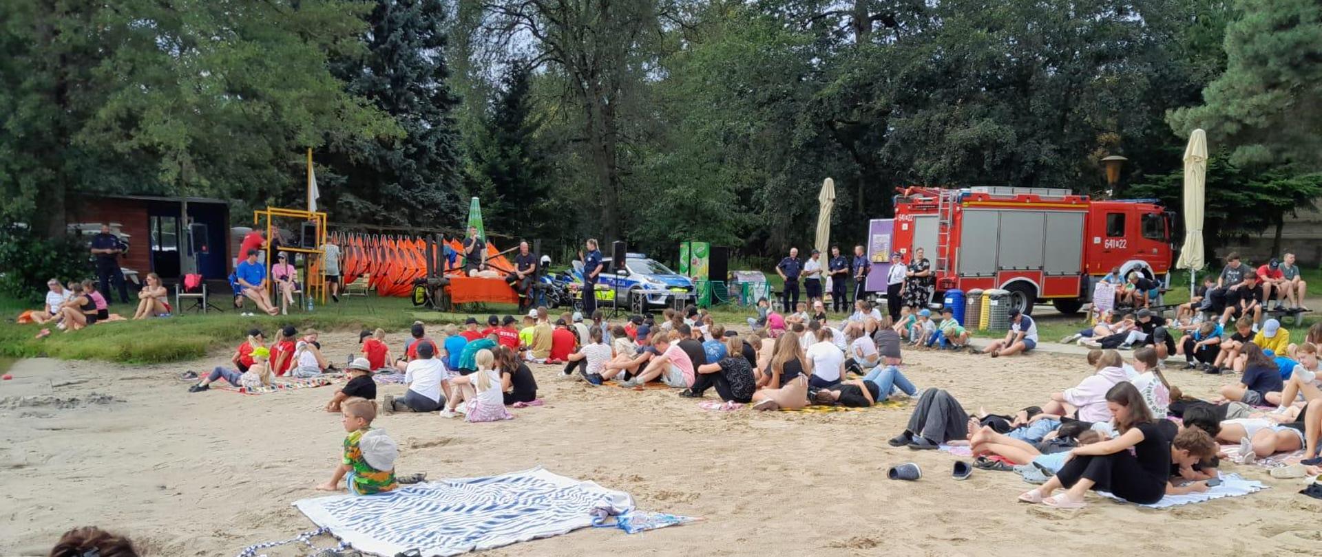 Zdjęcie wykonane na zewnątrz. Przedstawia uczestników spotkania podczas akcji „Kręci mnie bezpieczeństwo nad wodą 2024”. W tle widać wóz strażacki oraz wozy policyjne.