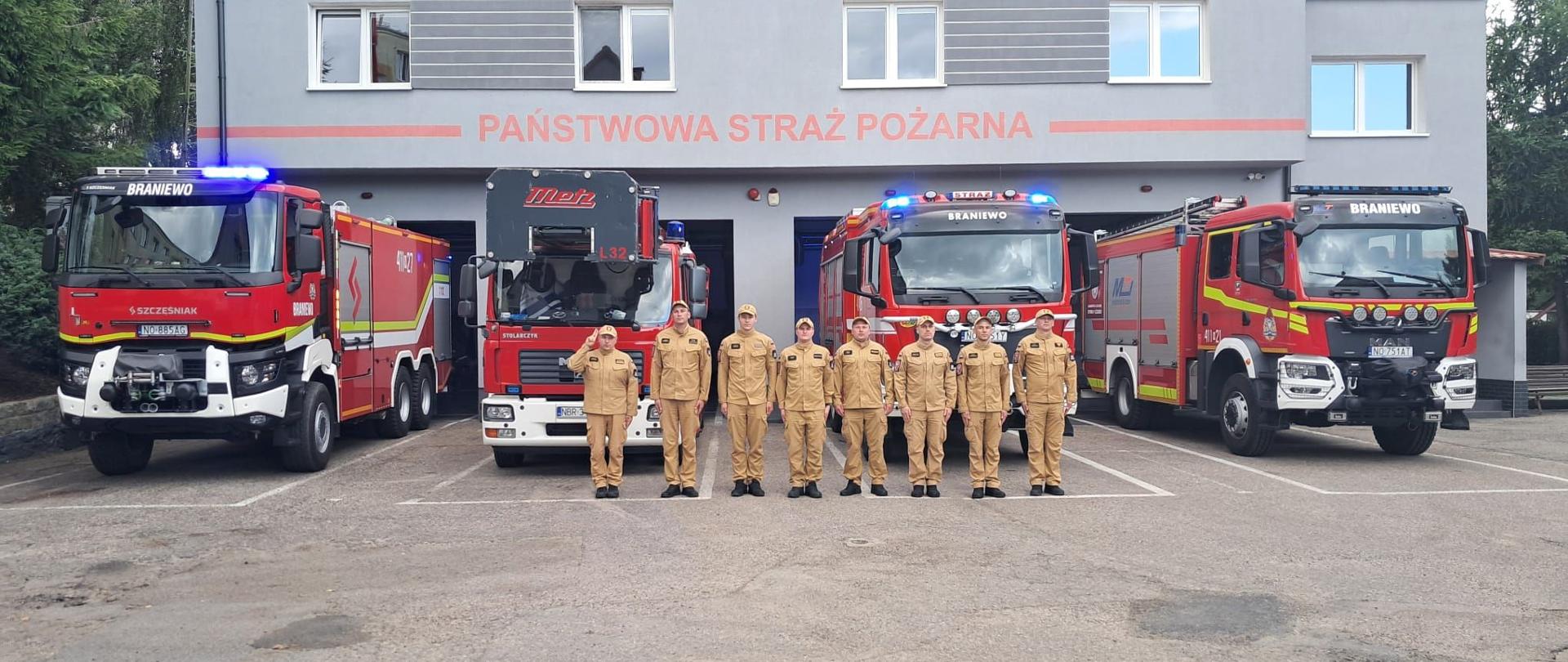Strażacy w jasnych mundurach na tle czterech samochodów strażackich i remizy.