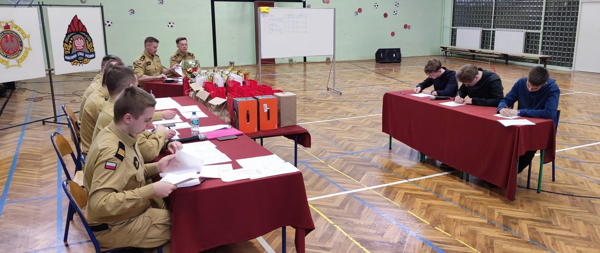 2. Na zdjęciu widać komisję sędziowską w skład której wchodzą funkcjonariusze Państwowej Straży Pożarnej oraz młodzież biorącą udział w turnieju podczas eliminacji ustnych. Miejscem przeprowadzania eliminacji jest sala gimnastyczna.