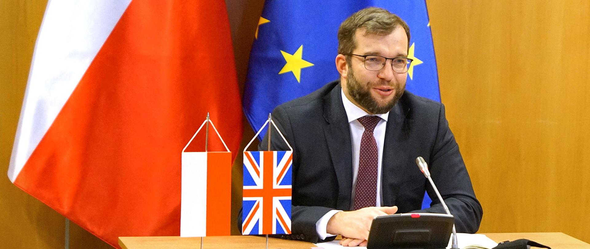 Minister Grzegorz Puda siedzący na tle flagi polskiej i Unii Europejskiej
