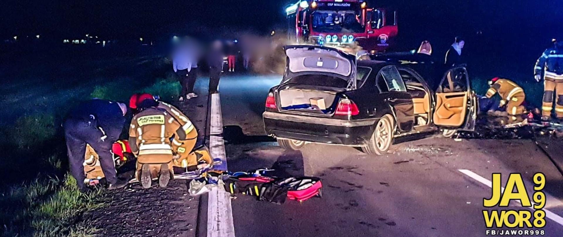 Obraz przedstawia strażaków udzielających pomocy poszkodowanemu w wypadku. Na środku jezdni uszkodzony pojazd osobowy. W tle pojazd pożarniczy i osoby postronne.