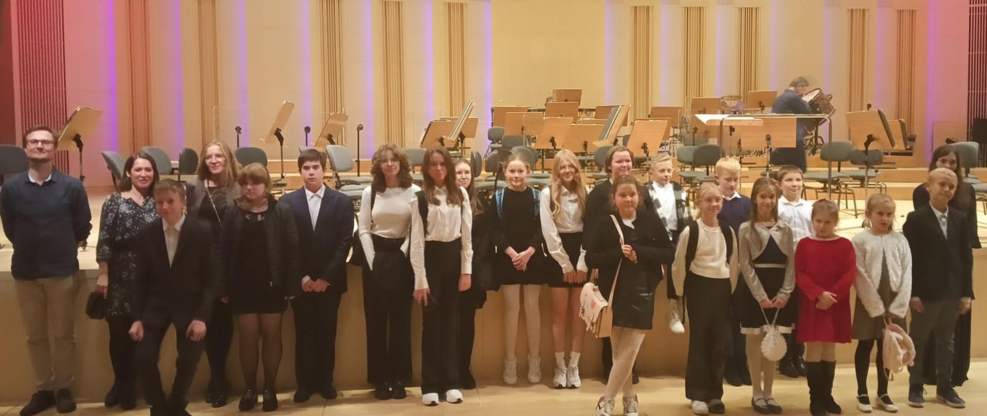 Na zdjęciu widać dzieci i nauczycieli stojących przed sceną w filharmonii.