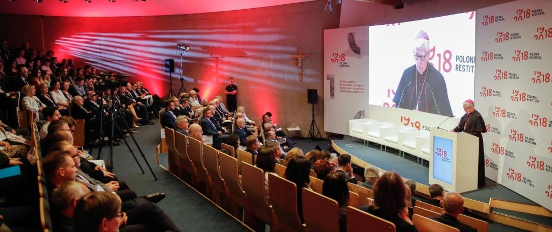 Biskup przemawia na tle ściany z logo Polonia Restituta