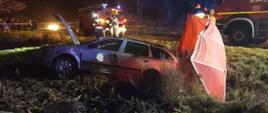 Zdjęcie przedstawia samochód osobowy marki Skoda koloru srebrnego, znajdujący się w przydrożnym rowie z otwartą maską oraz parawan. Ponadto na zdjęciu znajdują się samochody ratowniczo - gaśnicze oraz strażacy. 
