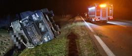 Kolizja na drodze krajowej. Auto ciężarowe leży w rowie na swoim prawym boku. Na jezdni stoi wóz straży pożarnej. Pora nocna. teren oświetlany jest a auta pożarniczego.