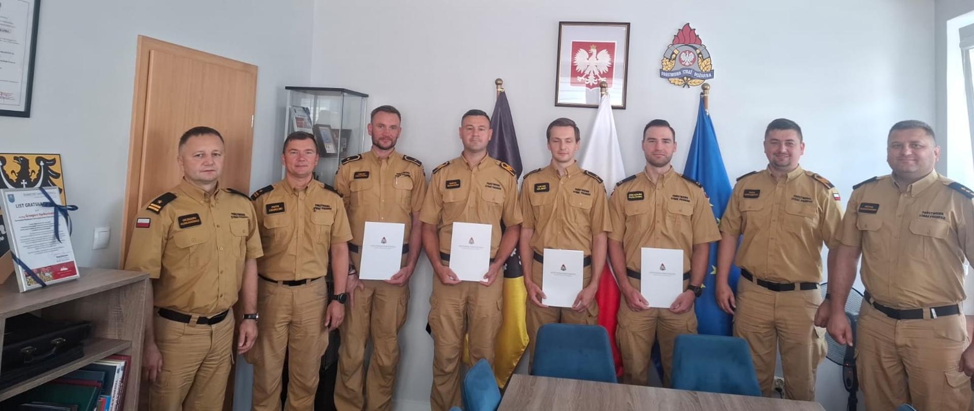 Ośmiu strażaków ubranych w żółte mundury, czterech z nich trzyma białe teczki z dokumentami z logo PSP. 