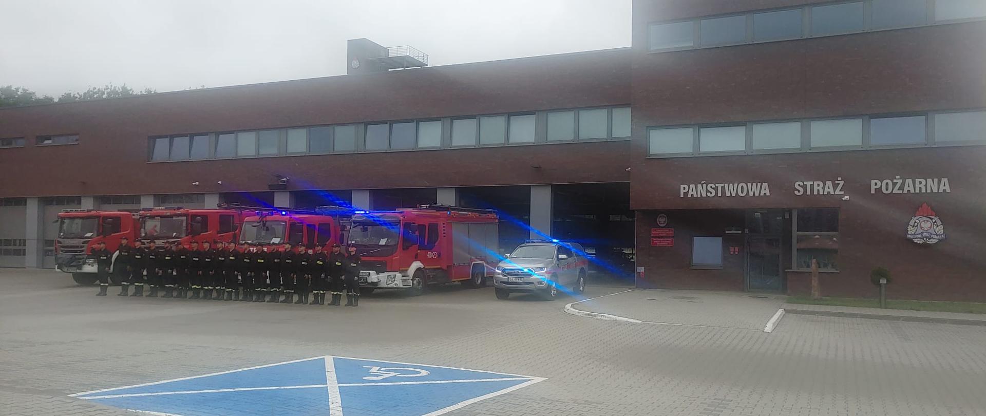 Na zdjęciu strażacy ustawieni w szeregu, za strażakami ustawione wozy bojowe w tle widoczny budynek Komendy Miejskiej Państwowej Straży Pożarnej w Bytomiu.