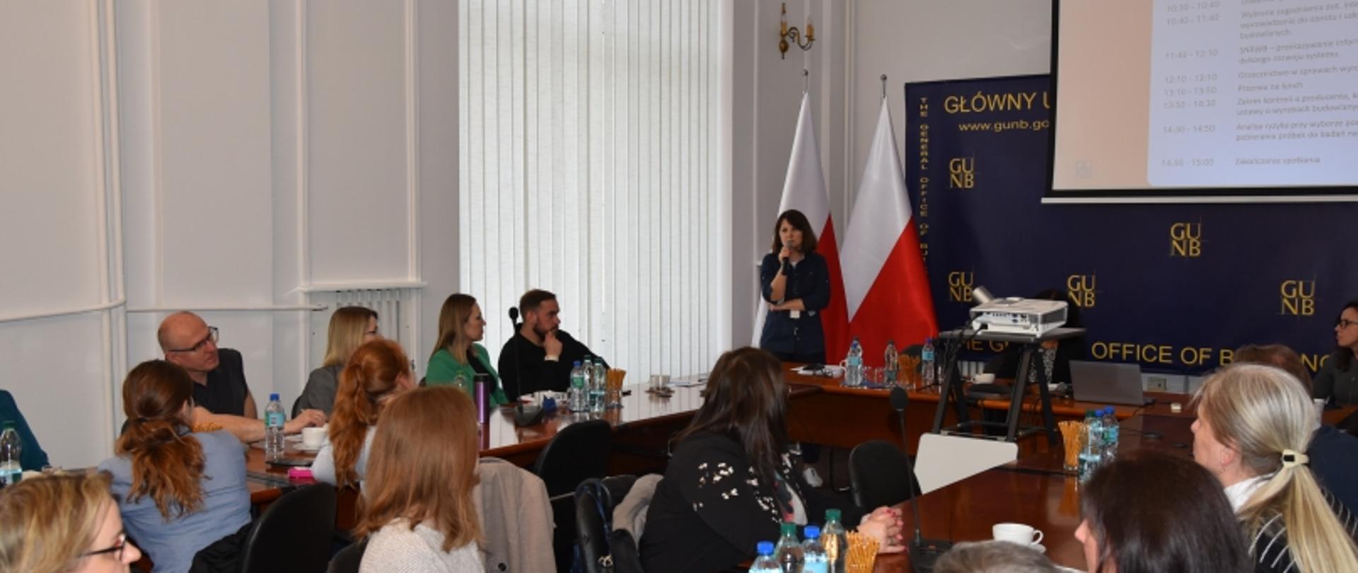 Prezentacja podczas spotkania w GUNB; uczestnicy przy stole, prelegent przy ekranie, w tle flagi i baner z adresem www.gunb.gov.pl.