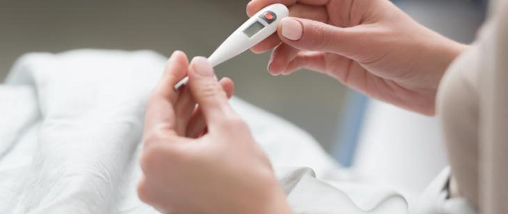 Na zdjęciu dwie kobiece dłonie trzymają elektryczny termometr.