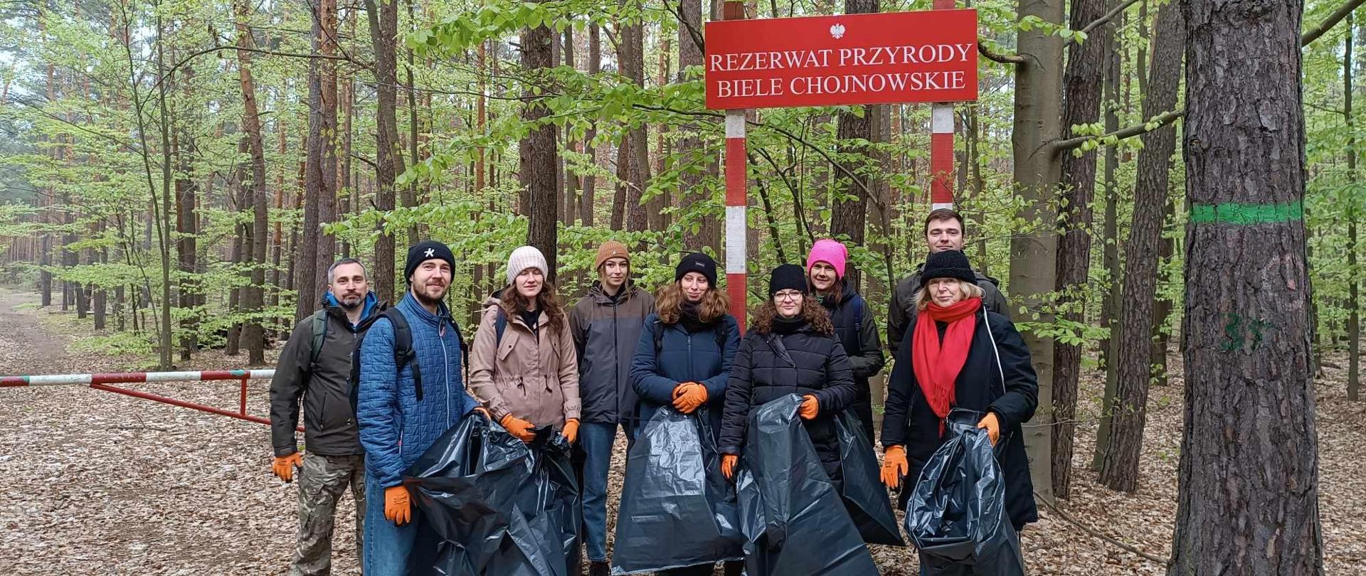 Grupa osób z workami na śmieci, w rękawiczkach stoi pod tablicą z nazwą rezerwatu Biele Chojnowskie, przed szlabanem prowadzącym do lasu 