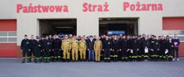 Plac zewnętrzny przed budynkiem Komendy Powiatowej PSP w Rawiczu. Do wspólnego zdjęcia pozują uczestnicy szkolenia wraz z zastępcą komendanta powiatowego PSP oraz instruktora prowadzącego zajęcia. Otwarte są dwie bramy garażowe, w kórych stoją wozy bojowe. Nad bramami widnieje napis "Państwowa Straż Pożarna"