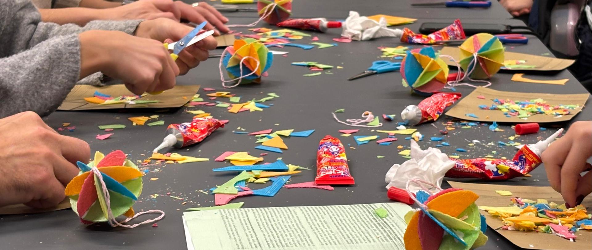 Warsztaty tworzenia tradycyjnych ozdób świątecznych „światy” z kolorowego opłatka; na stole leżą kleje, nożyczki i gotowe wielobarwne kule.