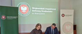 Przy stole na sali konferencyjnej siedzą Mazowiecki Wojewódzki Inspektor Ochrony Środowiska Krzysztof Cząstkiewicz i Mazowiecki Wojewódzki Lekarz Weterynarii Izabela Floryszczyk. Przedstawiciele obu inspekcji podpisują leżące przed nimi kopie porozumienia.